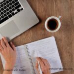 The corner of a laptop, a cup of coffee, and a person's hands writing in a notebook a viewed from overhead. Image represents continued learning.